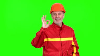 Kırmızı üniformalı mutlu gülen inşaat işçisi iyi niyet işareti veriyor. Yeşilde izole edilmiş.