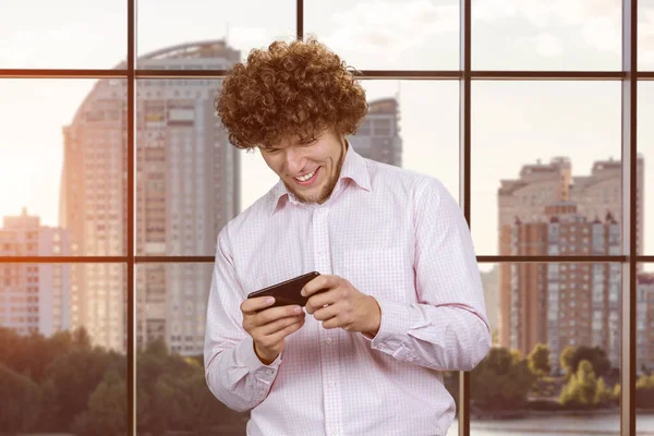 Excited happy young caucasian man gamer playing using smartphone. Guy playing mobile videogame app or watching a video stream holding cell phone indoors.