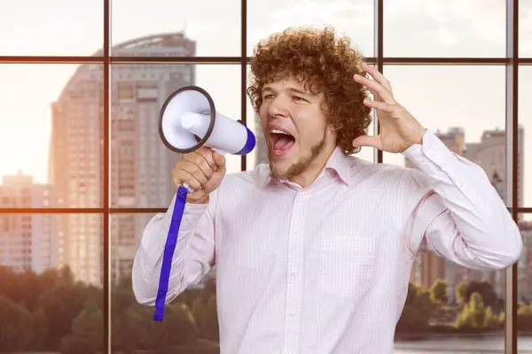 Portrait of a young excited expressive caucasian man with curly hair in white shirt screaming hot news shout in megaphone. Indoor window in the background.