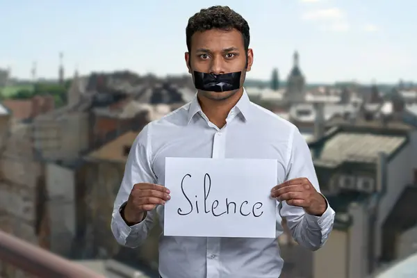 タップされた口を持つインド人男性は 碑文沈黙のポスターを保持しています バックグラウンドの輝く都市の建物 — ストック写真