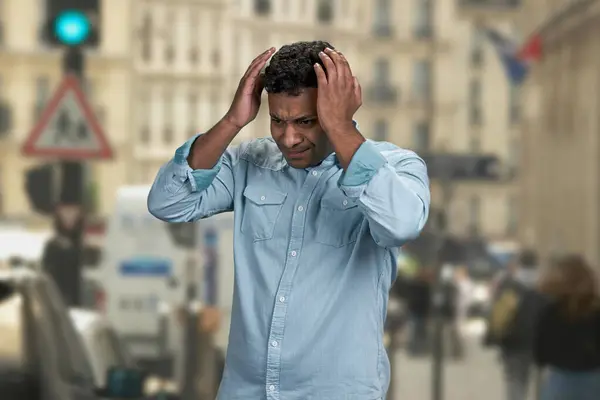 Stressed young man holding hands on head because of terrible headache. Blur crowded city street in the background.