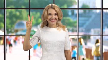 Neşeli sarışın, gülümseyen olgun bir kadın içeride durup zafer işareti veriyor. Arka planda yürüyen insanlar olan kapalı pencere..