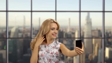 İçeride poz veren ve selfie çeken mutlu sarışın bir kadının portresi. Arkaplanda şehir manzarası olan işaretlenmiş pencere.