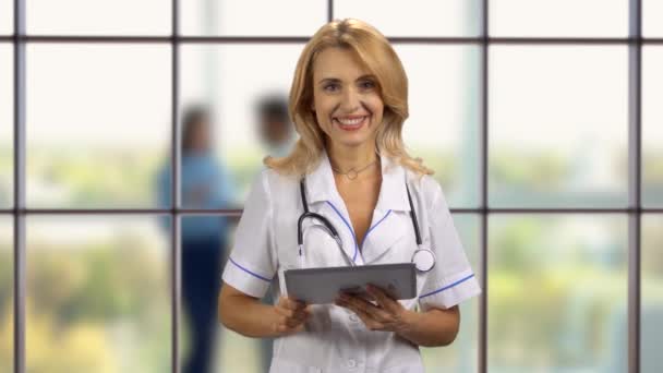 タブレットデバイスで立っている幸せなブロンドの女性医師の肖像画 バックグラウンドの室内窓 — ストック動画