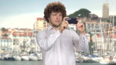 Kıvırcık saçlı genç bir turist akıllı telefonundaki fotoğraf makinesiyle video çekiyor. Arka plandaki yat rıhtımları.