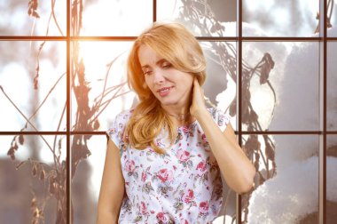 Portrait of mature blonde woman touching her neck because of pain. Indoor window in the background. clipart