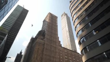 Midtown Manhattan, New York 'taki çeşitli gökdelenlerin çarpıcı manzarası. Binalar modern cam cepheleri, tarihi tasarımları ve parlak mavi gökyüzünün altında devam eden yapıları sergiliyor..