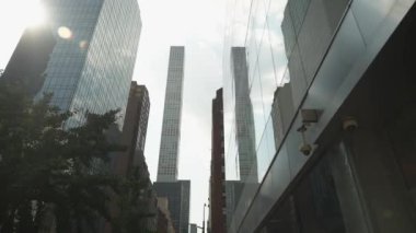 Camdan yansımalar ve şehir yeşiliyle çevrili gökdelenlerin parlak bir gökyüzüne karşı yükselişinin dramatik bir görüntüsü. Görüntü modern mimariyi ve kentsel canlılığı yakalıyor..