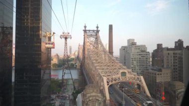 Bu görüntü, Roosevelt Adası Tramvayı 'nın yanından geçtiği Doğu Nehri boyunca uzanan ikonik Queensboro Köprüsü' nü vurgular. Kentsel ufuk çizgisi ve nehir çarpıcı bir şehir manzarası yaratır..
