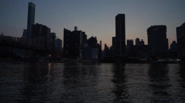 Manhattan silueti ve Queensboro Köprüsü güneş batarken aydınlanıyor. Akşamüstü gökyüzünün altındaki Doğu Nehri 'nin sakin sularında yansımalar dalgalanır..