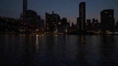 Queensboro Köprüsü gece New York şehrinin siluetinin arka planında parlar. Şehir ışıklarının yansımaları sakin nehirde parıldıyor, sakin bir şehir manzarası yaratıyor..