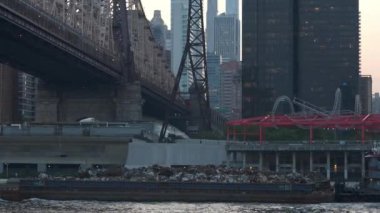 Gün doğumunda Queensboro Köprüsü 'nün altında metal parçaları taşıyan bir mavna yüzer. Kentsel arkaplanda gökdelenler, sanayi yapıları ve ikonik Roosevelt Adası tramvayı yer alıyor..
