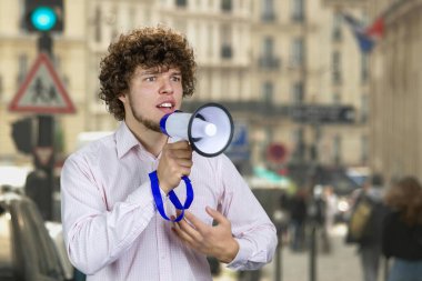 Portrait of a handsome young man with curly hair in white shirt speaks in loudspeaker. City street in the background. clipart