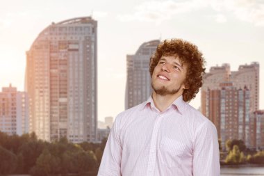 Happy joyful smiling young man with curly hair looking up. Thinking of new good opportunities, dreaming. Residential area buildings in the background. clipart