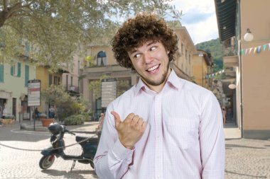 Baş parmağıyla arkasını gösteren kıvırcık saçlı mutlu genç adamın portresi. Arka planda şehir sokağı.
