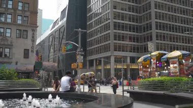 New York City, New York, ABD - 07.29.2024: Ziyaretçiler merkez bir çeşmenin yer aldığı canlı bir şehir meydanının keyfini çıkarırken, diğerleri yakınlardaki yiyecek tezgahlarını keşfediyorlar. Parlak güneş içerideki canlı atmosferi güçlendirir.