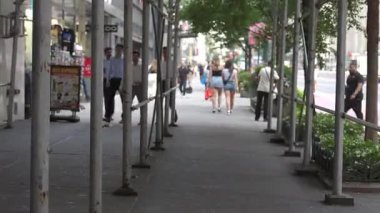 New York City, New York, ABD - 07.29.2024: Yayalar gölgeli bir şehir yolunda yürürken iskele ile kaplı bir şehir kaldırımı. Sahne günlük şehir yaşamını vitrinlerle, yeşilliklerle çekiyor.