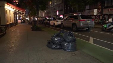 New York City, New York, ABD - 07.29.2024: Manhattan, New York 'ta, arabalar geçerken kaldırımda bir yığın çöp torbası duruyor. Sokak loş ışıklandırılmış ve kentsel atık yönetimini vurguluyor