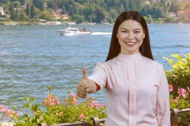 Genç gülümseyen Asyalı kadın başparmağını kaldırıyor. Açık hava doğa nehir arka planı.