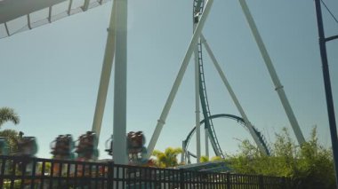 Bir lunapark treni açık mavi bir gökyüzüne doğru dik bir piste tırmanır ve arka planda güneş ışığı parıldar. Biniciler bu eğlence parkında coşkulu bir düşüş yaşamak üzereler..