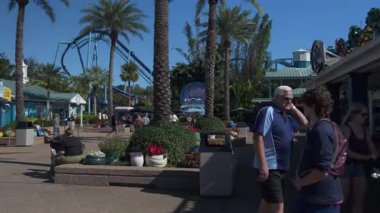 Orlando, Florida, ABD - 11.15.2024: Ziyaretçiler canlı bir deniz gösterisinde geziniyor, güneşli havanın ve arka planda heyecanlı yolculukların tadını çıkarıyorlar. Aileler parkın hareketli atmosferini emerken çeşitli dükkanları keşfediyorlar..