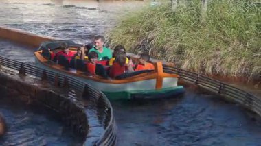 Orlando, Florida, ABD - 11.15.2024: Bir grup çocuk ve bir yetişkin bir tema parkındaki bir teknede oturuyorlar. Yol, otlar ve suyla çevrili yol boyunca yavaşça ilerliyor. Eğlenceli ve güvenli bir deneyim yaratıyor..