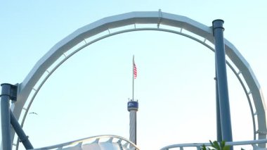 Birleşik Devletler bayrağı, açık mavi gökyüzündeki beyaz roller coaster pistinin dramatik çelik yayı tarafından çerçevelenmiş uzun bir gözlem kulesinin üzerinde dalgalanıyor. Bu çarpıcı kompozisyon bir eğlence parkı deneyiminin heyecan ve vatanseverlik atmosferini yakalar..