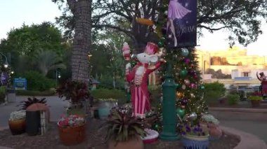 Orlando, Florida, ABD - 11.15.2024: Şenlikli giyinmiş bir fare heykeli, gün batımından hemen önce renkli süslemeler ve yemyeşil yapraklarla çevrili bir deniz parkında gururla duruyor.