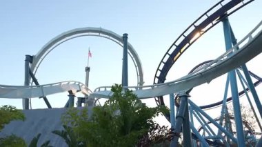 Güneş batarken gökyüzüne çarpıcı bir şekilde çarpan beyaz ve mavi hız treninin yakın görüntüsü. Güneş yapının içinde parıldıyor, döngü tasarımını vurguluyor. Amerikan bayrağı arka planda görünür, dinamik bir...