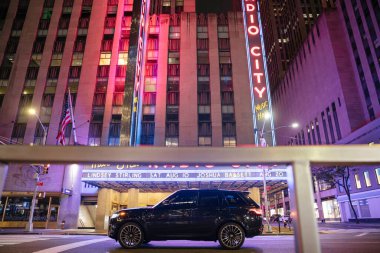 New York City, New York, ABD - 07.28.2024: New York 'taki Radio City Music Hall' un önünden siyah bir SUV geçer. Neon tabelalar pembe ve mavi parlıyor, gece hayatına renk katıyor..