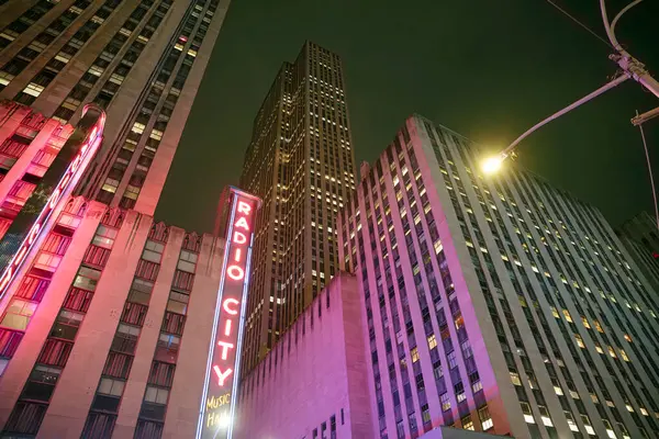 Manhattan 'ın hareketli caddelerinde kalabalıklar yürürken canlı neon ışıkları Radyo Şehri' ni aydınlatıyor. Gökdelenler gece gökyüzünün altında, dinamik bir şehir atmosferi yaratıyor..