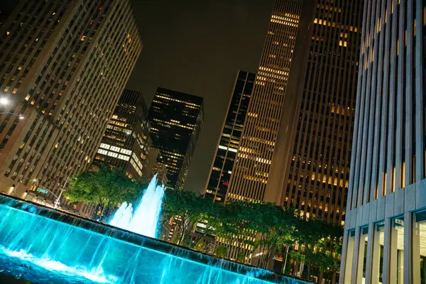 Manhattan 'da gece, parlak mavi bir çeşme yüksek gökdelenlerle çevrili yoğun bir meydanı aydınlatır. İnsanlar şehir ortamının hayat dolu atmosferinin tadını çıkararak geziniyor..