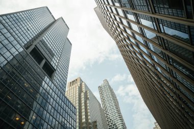 Camdan cepheli ve yansıtıcı pencereli modern yüksek katlı ofis binaları. Fotoğraf, şehir mimarisini gözler önüne seren parlak, hafif bulutlu bir gökyüzü altında dramatik bir şehir perspektifi yakalıyor..