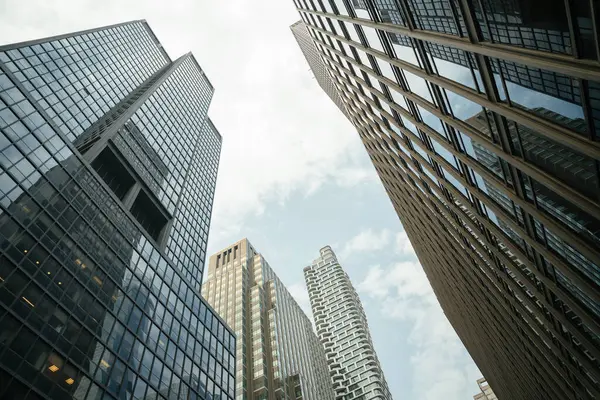 Camdan cepheli ve yansıtıcı pencereli modern yüksek katlı ofis binaları. Fotoğraf, şehir mimarisini gözler önüne seren parlak, hafif bulutlu bir gökyüzü altında dramatik bir şehir perspektifi yakalıyor..