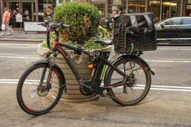Büyük bir kargo kutusu olan elektrikli bir bisiklet New York caddesinde bir saksının yanında park edilmiş. Sahne kentsel yaşamı, iş ekonomisini ve çevre dostu ulaşımı yansıtıyor.