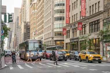 New York City, New York, ABD - 07.29.2024: Yayalar New York Halk Kütüphanesi yakınlarındaki kalabalık bir Manhattan caddesini geçtiler. Sarı taksiler, otobüsler ve bisikletçiler kırmızı kütüphane afişleri yukarıda asılı dururken şehir trafiğinde ilerliyorlar.