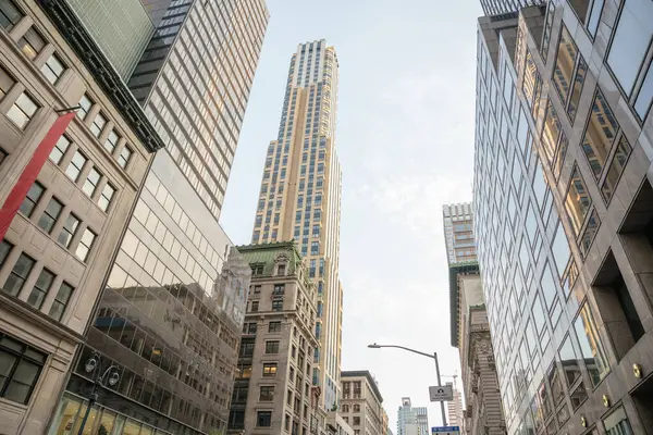 Manhattan şehir merkezindeki tarihi ve çağdaş binaların üzerinde modern bir cam gökdelen yükseliyor. Yansımalar, dikey çizgiler ve yumuşak sabah ışığı mimari karşıtlığı vurgular.