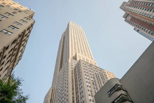Empire State Binası çevresindeki binaların üstünde. Alçak bir açıdan çekilen gökdelen, açık mavi gökyüzüne karşı görkemli görünüyor..
