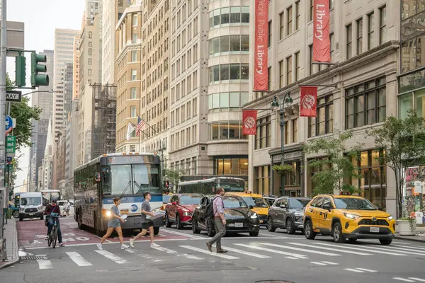 New York City, New York, ABD - 07.29.2024: Yayalar New York Halk Kütüphanesi yakınlarındaki kalabalık bir Manhattan caddesini geçtiler. Sarı taksiler, otobüsler ve bisikletçiler kırmızı kütüphane afişleri yukarıda asılı dururken şehir trafiğinde ilerliyorlar.