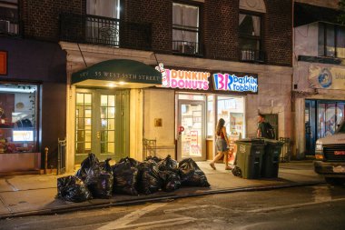New York City, New York, ABD - 07.29.2024: Çöp torbaları ve çöp bidonları kaldırıma dizilmiş, günlük şehir yaşamını yansıtıyor. Bir kadın gece vakti New York 'ta West 55. Cadde' deki Dunkin Donutları ve Baskin Robbins 'in yanından geçiyor..
