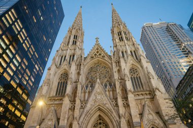 New York 'taki modern cam gökdelenler arasında yükselen St. Patricks Katedrali' nin ikonik Gotik Uyanış kuleleri. Süslü taş cephe ve detaylı vitray pencereler akşam ışığı sönerken göze çarpıyor..