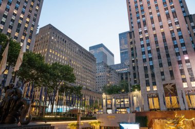 New York City, New York, ABD - 07.29.2024: New York 'taki Rockefeller Center' da yüksek binalar, sallanan bayraklar ve Prometheus heykelinin yer aldığı bir akşam sahnesi. Aydınlatılmış ofis pencereleri ve plaza ışıkları canlı bir şehir atmosferi yaratır.