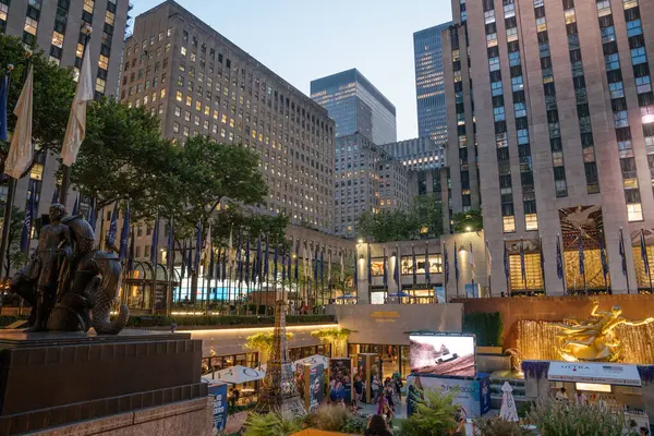 New York City, New York, ABD - 07.29.2024: Akşam ışıkları Rockefeller Plaza 'yı altın Prometheus heykeli ve uluslararası bayraklarla aydınlatıyor. Sahnede bir sergi alanı ve New York simgesinin tadını çıkaran ziyaretçiler yer alıyor..