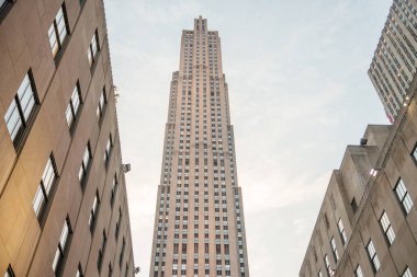 Art Deco binaları arasında yükselen Rockefeller Center kulesinin çarpıcı görüntüsü. Günbatımının yumuşak parıltısı taş cephesini aydınlatıyor, dikey simetriyi ve ikonik mimariyi vurguluyor..