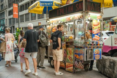 New York, New York, New York, ABD - 07.29.2024: New York 'ta bir helal gıda arabası kalabalık bir kaldırımda müşterilere sosisli sandviç, kraker ve içecek sunmaktadır. İnsanlar levhalar, şemsiyeler ve hareketlerle dolu canlı bir sokak sahnesinde bekler ve yürür..