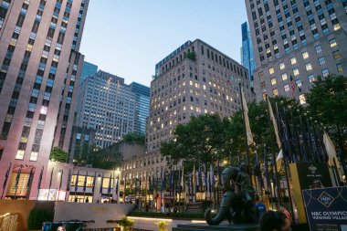 New York City, New York, ABD - 07.29.2024: New York 'taki Rockefeller Center' da NBC Olimpiyat Viewing Village için yüksek binalar ve bayraklar. İnsanlar, modern gökdelenler ve dekoratif heykellerle aydınlık meydanda yürüyorlar..