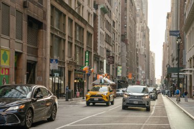 New York City, New York, ABD - 07.29.2024: Sarı bir taksi ve diğer arabalar Manhattan 'da dar bir cadde boyunca uzun binalarla çevrili. İnsanlar sıcak akşam ışığında şehir hareketlenirken kaldırımlarda yürür..