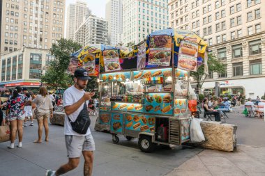 New York City, New York, ABD - 07.29.2024: Canlı bir New York şehir meydanında canlı bir helal gıda arabası insanlarla çevrilidir. Renkli tabelalarda sosisli, pirinç tabağı ve içecekler sergilenirken, yayalar açık hava ortamının keyfini çıkarıyorlar..