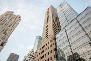Manhattan şehir merkezindeki modern yüksek katlı ofis binaları gökyüzüne ulaşıyor. Yansıtıcı cam pencereler mavi gökyüzü ve bulutları yansıtarak dinamik bir şehir manzarası yaratıyor..
