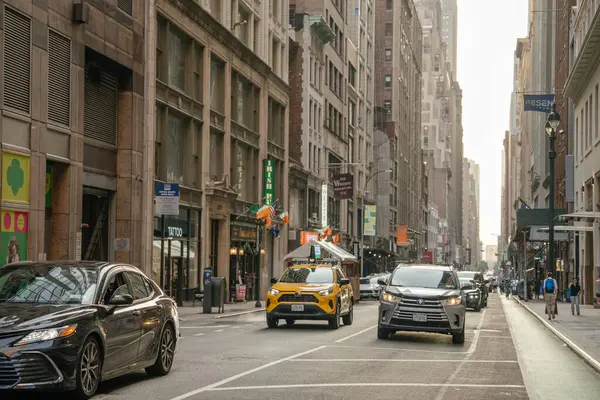 New York City, New York, ABD - 07.29.2024: Sarı bir taksi ve diğer arabalar Manhattan 'da dar bir cadde boyunca uzun binalarla çevrili. İnsanlar sıcak akşam ışığında şehir hareketlenirken kaldırımlarda yürür..